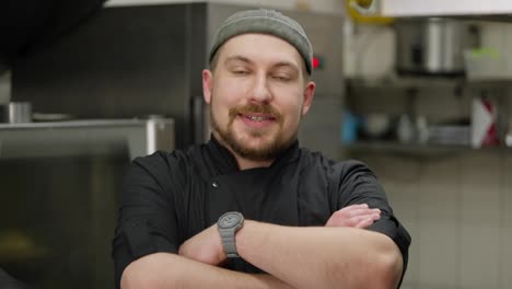 Retrato-De-Un-Chef-Confiado-Y-Feliz-Con-Barba-En-Un-Uniforme-Negro-En-La-Cocina-De-Un-Restaurante.-Jefe-Alegre-Con-Un-Uniforme-Negro-Posando-Y-Mirando-A-La-Cámara-Cruzando-Los-Brazos-Sobre-El-Pecho.