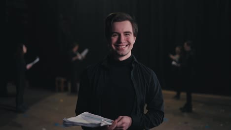 Portrait-of-a-happy-young-male-actor-in-a-black-suit-with-papers-in-his-hands-against-of-actors-who-are-rehearsing-for-a-performance-on-stage-in-a-theater-with-black-curtains