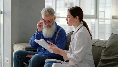 Un-Hombre-Mayor-De-Pelo-Gris-Y-Barba-Frondosa-Con-Gafas-Y-Camisa-Azul-Mira-Hojas-De-Papel-Que-Una-Médica-Le-Muestra-Durante-Una-Consulta-A-Domicilio-En-Un-Apartamento-Moderno.