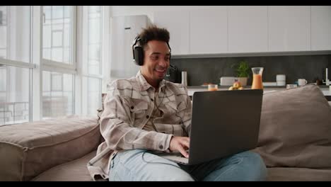 A-happy-Black-skinned-young-brunette-man-with-stubble-wearing-black-modern-headphones-and-a-cream-plaid-shirt-plays-a-video-game-on-his-gray-laptop-while-sitting-on-a-modern-sofa-in-a-modern-studio-apartment