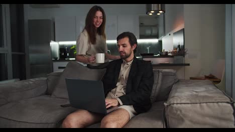 Happy-brunette-man-with-stubble-in-a-black-jacket-works-at-his-gray-laptop-while-his-brunette-girlfriend-in-a-beige-T-shirt-brings-him-tea-on-the-sofa-in-a-modern-studio-apartment-in-the-evening