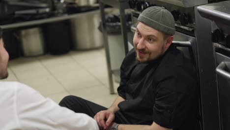 Close-up-over-the-shoulder-of-a-confident-and-happy-cook-with-a-beard-in-a-black-uniform-talking-to-a-waiter-in-a-white-shirt-while-sitting-on-the-floor-in-the-kitchen-in-a-restaurant