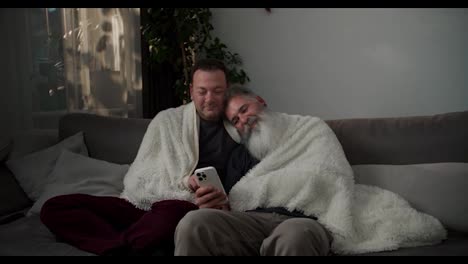 A-happy-brunette-man-sits-on-the-sofa-wrapped-in-a-white-woolen-blanket-with-his-elderly-boyfriend-with-gray-hair-and-a-lush-white-beard-they-are-looking-at-something-on-their-white-smartphone-on-a-gray-sofa-in-a-modern-apartment