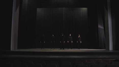 A-group-of-actors-in-black-stage-clothes-in-the-theater-perform-synchronously-and-practice-synchronized-movement-in-on-stage-in-the-theater-near-black-curtains