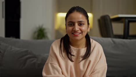 Portrait-of-a-happy-brunette-girl-with-brown-skin-in-a-beige-jacket-who-sits-on-a-gray-sofa-in-a-modern-apartment-during-the-day.-Portrait-of-a-happy-brunette-girl-in-a-modern-apartment-during-the-day