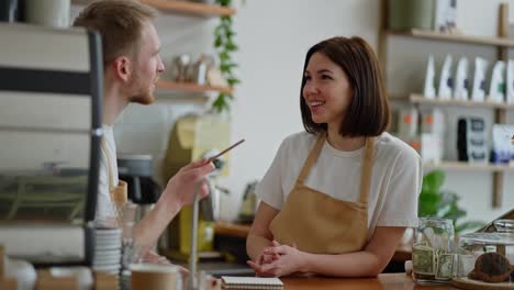 Happy-brunette-girl-in-a-white-T-shirt-a-cafe-worker-stands-behind-the-counter-talking-to-a-guy-barista-while-drawing-up-his-plan-in-a-cafe