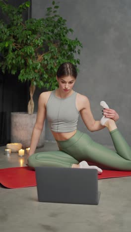 Vertical-video-of-a-confident-brunette-girl-in-a-sports-top-and-green-pants-sitting-on-a-red-mat-and-doing-sports-and-stretching-with-the-help-of-online-lessons-through-her-gray-Laptop-at-home-in-a-modern-apartment-near-burnt-candles-and-a-large-indoor-plant