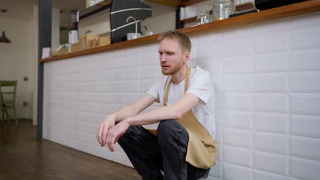 Tired-cafe-worker-waiter-sitting-near-a-white-wall-and-trying-to-take-a-break-exhaling-and-massaging-his-head-during-a-hard-day-at-work