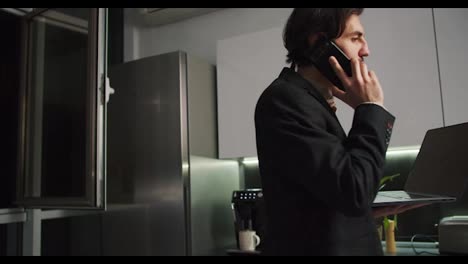 A-brunette-man-with-stubble-in-a-black-jacket-walks-around-the-kitchen-with-a-laptop-in-his-hands-and-talks-on-the-phone-without-pants-and-only-in-shorts-during-a-video-conference-and-working-remotely-in-a-modern-apartment-in-the-evening