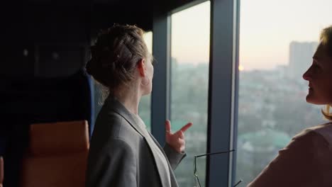 A-confident-middle-aged-businesswoman-in-a-gray-business-suit-communicates-with-her-colleague-while-standing-near-panoramic-windows-overlooking-the-city-in-a-modern-office