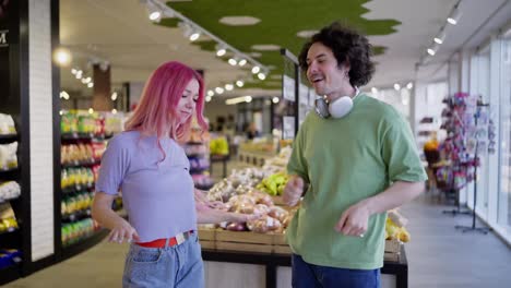 Happy-girl-with-pink-hair-and-brunette-guy-in-a-green-T-shirt-dancing-in-a-grocery-store.-Happy-bright-couple-dance-in-grocery-store-while-shopping