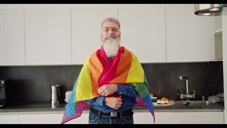 Ein-Glücklicher-älterer-Mann-Mit-Grauem-Haar-Und-üppigem-Bart-Und-Brille-Erhebt-Sich-über-Sich-Und-Umarmt-Dann-Eine-LGBT-Person-In-Einer-Modernen-Küche-In-Einer-Sonnigen-Wohnung.-Porträt-Eines-Glücklichen-älteren-Mannes,-Der-Die-LGBT-Gemeinschaft-Unterstützt-Und-Neben-Der-LGBT-Flagge-Posiert