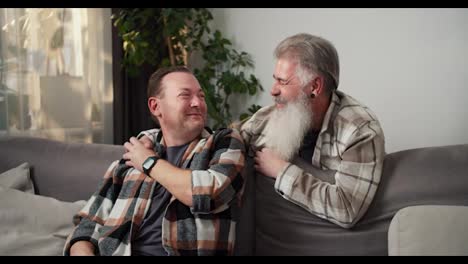 Happy-elderly-man-with-gray-hair-and-a-lush-beard-in-a-checkered-shirt-communicates-with-his-brunette-boyfriend-and-holds-hands-with-him-while-hanging-out-at-home-near-the-sofa-in-a-modern-apartment