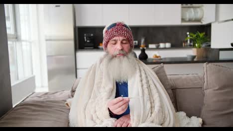 An-elderly-man-with-a-gray-beard-is-wrapped-in-a-warm-white-blanket-and-checks-his-temperature-with-a-thermometer.-He-sits-in-a-hat-and-feels-that-he-has-a-cold-in-a-modern-apartment