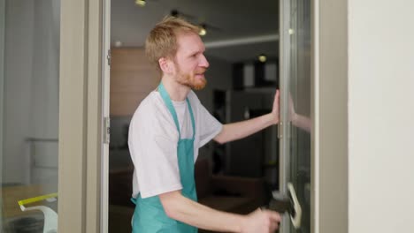 Side-view-of-a-confident-blond-male-cleaner-in-a-white-t-shirt-with-blue-aprons-cleaning-a-glass-wall-using-a-vacuum-cleaner-for-windows-in-a-modern-apartment