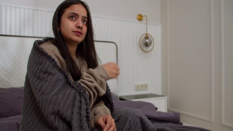 Sad-woman-with-a-cold-in-a-brown-jacket-and-a-dark-gray-stove-takes-pills-during-her-illness-Sitting-on-the-bed-in-a-modern-apartment-in-the-bedroom