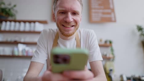 Close-up-of-a-happy-blond-guy-with-a-beard-in-a-white-T-shirt-looking-at-social-networks-using-a-green-phone-leaning-on-the-counter-in-a-cafe