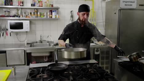 Confident-male-chef-in-a-black-uniform-and-with-protective-rubber-gloves-puts-a-saucepan-with-water-on-a-gas-stove-waiting-for-the-water-to-boil-in-a-modern-kitchen-in-a-restaurant