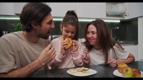 A-brunette-girl-in-pink-clothes-together-with-her-brunette-mother-in-a-white-T-shirt-and-a-brunette-father-with-stubble-in-a-beige-T-shirt-and-having-breakfast-in-the-morning-at-the-dining-table-in-a-modern-apartment-in-the-kitchen