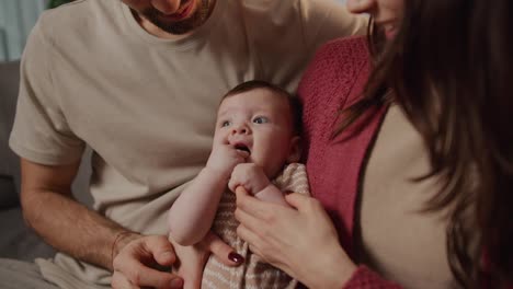 Primer-Plano-De-Una-Niñita-Con-Una-Camiseta-Marrón-Recostada-En-Los-Brazos-De-Su-Joven-Madre-Y-Mirando-A-Su-Alrededor-Mientras-Una-Familia-Joven-Pasa-Tiempo-Junta-En-Un-Apartamento-Moderno.