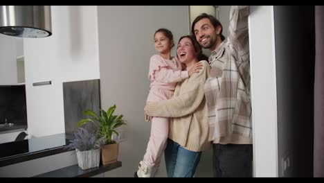 A-happy-brunette-girl-in-a-beige-sweater-carries-her-daughter-in-pink-clothes-in-her-arms-and-together-with-her-boyfriend-enters-a-modern-apartment-after-moving.-A-happy-family-enters-a-new-apartment-after-moving-in