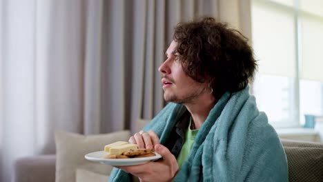 Happy-interested-brunette-guy-with-curly-hair-eats-sweet-snacks-and-watches-incredible-movies-while-sitting-on-the-couch-at-home-wrapped-in-a-blanket