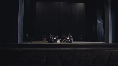 A-group-of-confident-young-actors-in-black-suits-together-with-their-stage-director-are-sitting-on-stage-and-preparing-for-their-next-performance-sorting-through-sheets-of-script-on-stage