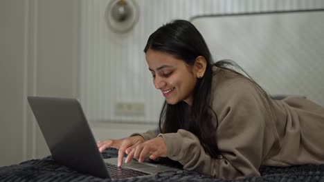 Ein-Glückliches,-Braunhäutiges,-Brünettes-Mädchen-In-Einem-Grauen-Pullover-Liegt-Auf-Dem-Bett-Und-Tippt-Auf-Ihrem-Laptop,-Während-Sie-In-Einem-Modernen-Apartment-Im-Schlafzimmer-Arbeitet-Und-Zeit-Online-Verbringt