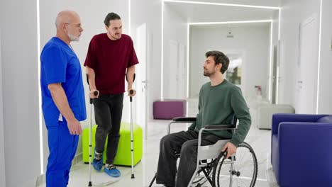 A-brunette-man-in-a-green-sweater-sits-on-a-wheelchair-near-a-man-who-stands-on-crutches-and-talks-with-the-doctor-about-their-injuries-and-the-duration-of-rehabilitation-in-a-modern-clinic