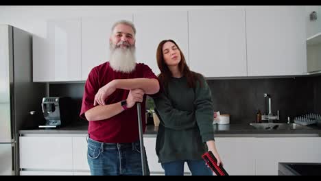 Portrait-of-a-happy-elderly-man-with-gray-hair-and-a-lush-beard-in-a-red-T-shirt-who-poses-with-his-adult-brunette-daughter-in-a-green-sweater-while-cleaning-in-a-modern-kitchen