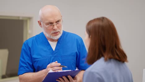 A-confident-man-with-glasses-an-elderly-doctor-in-a-blue-uniform-holds-a-blue-tablet-in-his-hands-and-talks-with-a-brunette-girl-about-her-problems-of-disease-prevention-and-diagnoses-in-a-modern-clinic