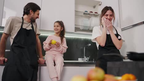 Happy-brunette-girl-talking-on-the-phone-in-a-white-T-shirt-and-black-apron-together-with-her-brunette-husband-with-stubble-communicate-and-have-fun-with-their-little-daughter-in-pink-clothes-while-preparing-for-breakfast-in-the-morning