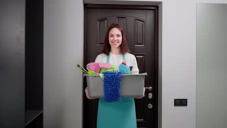 Portrait-of-a-confident-young-brunette-cleaning-lady-in-a-blue-apron-who-holds-in-her-hands-a-gray-basin-with-various-cleaners-and-items-for-cleaning-the-apartment