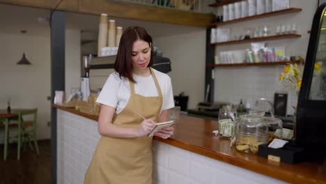 Portrait-of-a-brunette-waitress-girl-in-a-yellow-apron-who-makes-notes-in-her-notebook-near-the-cash-register-in-a-cafe