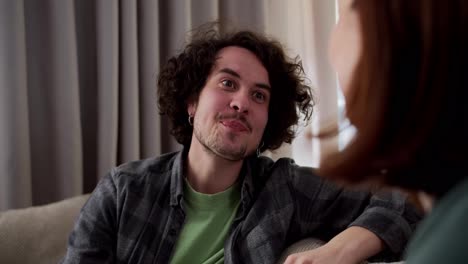 Close-up-over-the-shoulder-a-happy-brunette-guy-with-curly-hair-in-a-mustache-and-in-a-checkered-gray-shirt-communicates-with-his-brunette-girlfriend-while-sitting-on-the-sofa-at-home