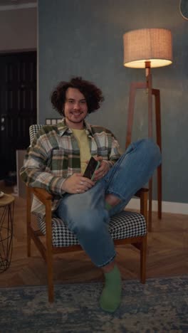 Vertical-video-portraits-on-the-left-of-a-brunette-guy-with-curly-hair-in-a-checkered-shirt-and-jeans-who-sits-on-a-chair-near-a-lamp-with-soft-light-in-a-modern-apartment-at-home