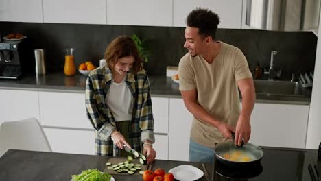 Happy-young-Black-skinned-brunette-man-in-a-cream-t-shirt-cooks-scrambled-eggs-and-frying-his-breakfast-while-his-mature-young-girlfriend-prepares-salad-in-the-morning-at-a-modern-apartment-on-a-black-kitchen-table