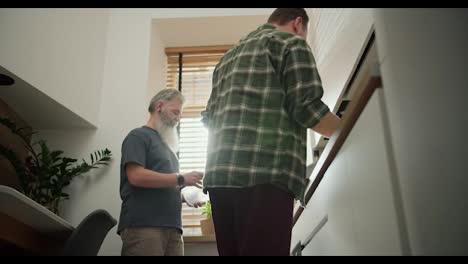 A-brunette-man-in-a-checkered-green-shirt,-together-with-his-elderly-boyfriend-with-gray-hair-and-a-lush-gray-beard-in-a-gray-T-shirt-are-cleaning-up-the-kitchen-after-preparing-lunch-together