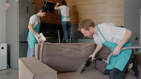 Side-view-of-a-confident-trio-group-of-cleaners-in-white-T-shirts-and-blue-aprons-wiping-down-an-apartment-and-a-man-using-a-vacuum-cleaner-cleaning-a-gray-sofa-in-a-modern-apartment-cleaning-on-call