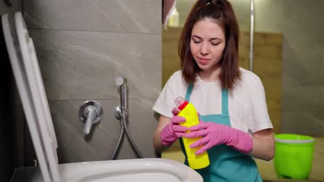 A-confident-brunette-cleaning-lady-in-a-white-T-shirt-and-blue-apron-sprays-detergent-from-a-yellow-bottle-on-the-walls-of-the-toilet-during-cleaning-in-the-bathroom-in-an-apartment-on-call