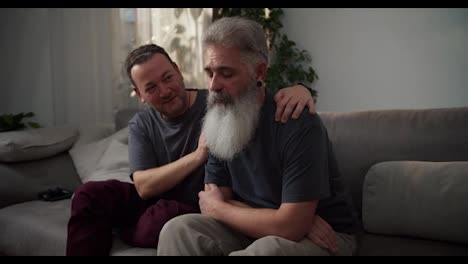 A-sad-man-with-gray-hair-and-a-lush-beard-with-a-black-earring-in-his-ear-and-in-a-gray-T-shirt-is-sad-and-his-middle-aged-brunette-boyfriend-supports-him-Sitting-on-the-sofa-in-a-modern-apartment-in-the-evening