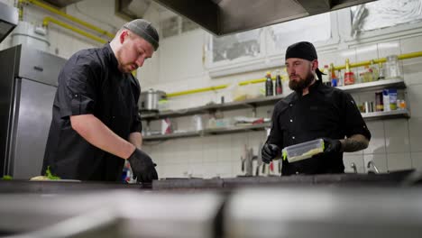 Two-chefs-in-black-uniforms-prepare-a-dish-and-share-their-experience-near-the-stove-in-the-kitchen-in-a-restaurant.-A-male-chef-with-a-beard-in-a-black-uniform-prepares-a-dish-under-the-supervision-of-a-more-experienced-chef-in-the-kitchen