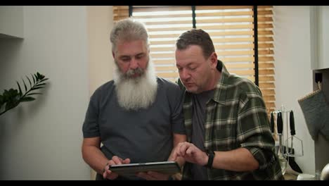 An-interested-middle-aged-brunette-man-with-stubble-in-a-green-checkered-shirt-together-with-his-elderly-boyfriend-with-a-gray-gray-beard-and-a-lush-mustache-are-choosing-something-on-their-electronic-tablet-during-the-day-at-home