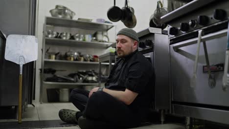 Portrait-of-a-very-tired-man-with-a-beard-of-a-professional-chef-in-a-black-uniform-who-sits-on-the-floor-leaning-on-the-table-in-the-kitchen-of-a-restaurant-and-sighs-heavily