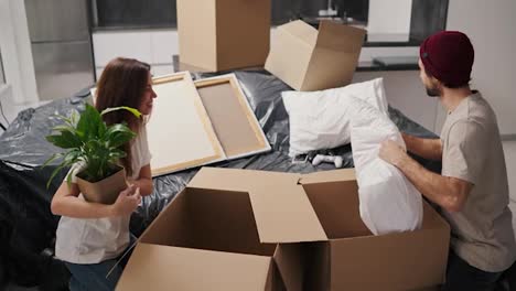 Happy-brunette-girl-in-a-white-T-shirt-holds-a-house-plant-in-her-hands-and-takes-items-out-of-a-box-together-with-her-boyfriend-in-a-beige-T-shirt-during-her-move-to-a-new-apartment