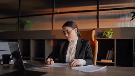 Zoom-in-portrait-of-a-confident-concentrated-brunette-girl-in-round-glasses-and-a-business-uniform-who-sits-at-a-wooden-table-in-the-office-in-front-of-a-laptop-and-writes-down-her-ideas-on-paper-while-working-in-a-sunny-office