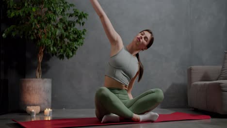 A-confident-and-calm-brunette-girl-in-a-summer-sports-uniform-sits-in-the-lotus-position-on-the-red-carpet-and-does-sports-and-stretching-near-lit-candles-and-a-houseplant-in-a-modern-apartment