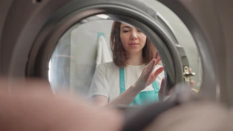 Blick-Von-Innen-Auf-Die-Waschmaschine-Porträt-Eines-Glücklichen-Brünetten-Mädchens-In-Einem-Weißen-T-Shirt,-Das-In-Einer-Modernen-Wohnung-Dinge-In-Die-Waschmaschine-Lädt-Und-Von-Außen-Hineinschaut