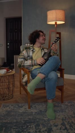 Vertical-video-of-a-happy-guy-with-curly-hair-in-a-plaid-shirt-sitting-on-a-chair-and-playing-a-ukulele-stringed-musical-instrument-at-home-in-the-evening