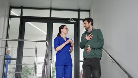 Una-Chica-Morena-Segura-De-Sí-Misma-Con-Gafas-Redondas,-Un-Médico-Con-Uniforme-Azul-Se-Comunica-Con-Un-Chico-Joven-Moreno-Con-Barba-Incipiente-Con-Una-Chaqueta-Verde-Y-Auriculares-Inalámbricos-Sobre-Sus-Síntomas-En-Una-Clínica-Moderna